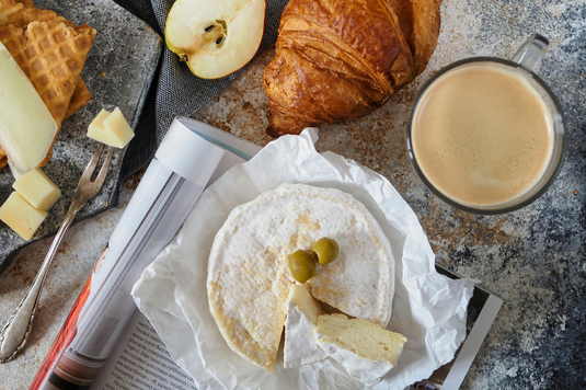 朝食の光景。ページが広げられた雑誌の上にカマンベールチーズのパッケージ。クロワッサン、りんご、バターが添えられたワッフル。コーヒーの入ったマグカップ。