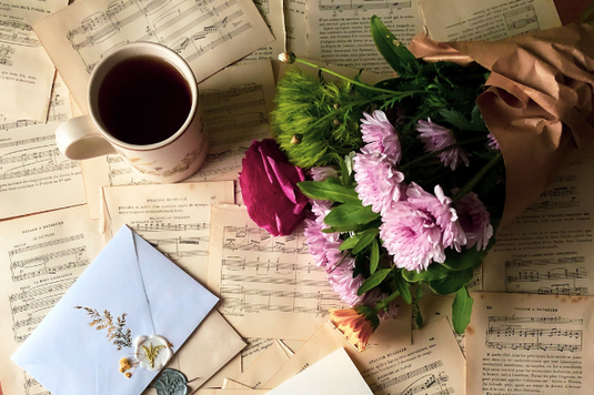テーブル一面に広げられた楽譜と封筒。その上に置かれた花束とコーヒーのマグカップ。