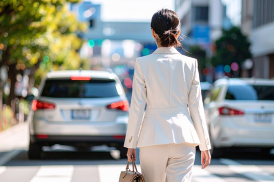 ビジネスバッグを下げ、スーツ姿で街を歩くビジネスパーソン。ベージュのパンツスーツの女性の後ろ姿。