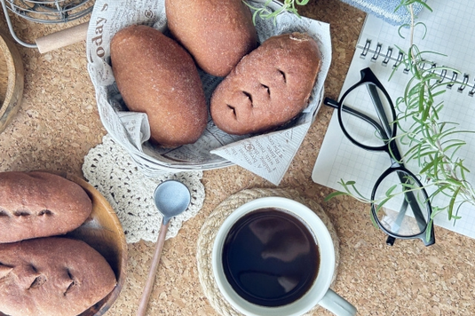 ページがめくられたリングメモ。黒縁眼鏡。チョコパンが盛り付けられたお皿。コーヒーの入ったマグカップ。