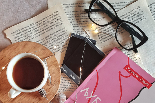 英文の資料ペーパーの上に置かれた黒ぶち眼鏡、ポラロイド写真、ピンク色の背表紙のペーパーバックス、コーヒーの入ったマグカップ。
