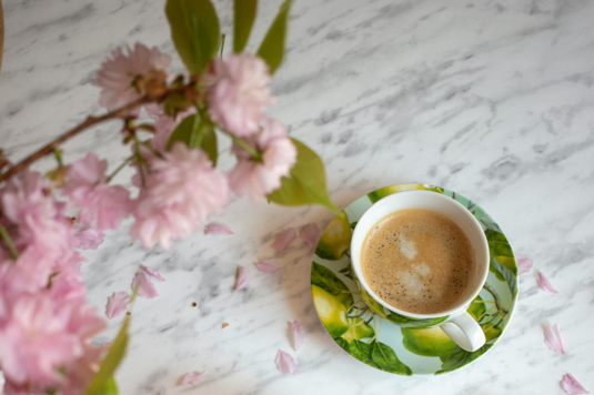 マーブル調のテーブルに置かれたコーヒーの入ったカップ＆ソーサ。花瓶に生けられた桜の小枝から花びらが散っている。