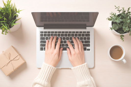 ノートパソコンのキーボードを叩く女性の指先。隣にギフトボックス。グリーンの鉢植。コーヒーの入ったマグカップ。