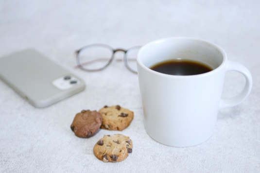 コーヒーの入ったマグカップ、スマートフォン、眼鏡、チョコチップクッキー。