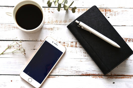 手帳の上に置かれたボールペン。スマートフォン。コーヒーの入ったマグカップ。