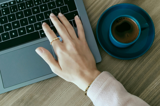 ノートパソコンのキーボードを叩くファッションリングをはめた女性の指先。コーヒーの入ったマグカップ。