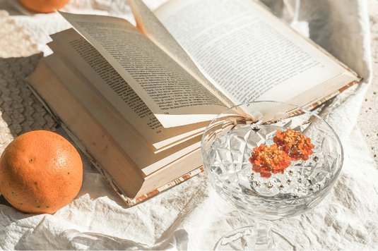 ページがめくられた本の傍らにオレンジが置かれている。オレンジ色の小さな花が浮かべられた水のグラス。