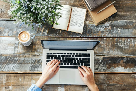 木製のテーブルに本とノートパソコンを広げて。キーボードを操作する女性の指先。傍らにコーヒーカップと花が生けられた花瓶。