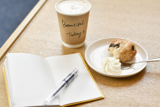 カフェのテーブルに広げられたノートとボールペン。チョコチップのスコーンと生クリームののったお皿とフォーク。紙コップのコーヒー。
