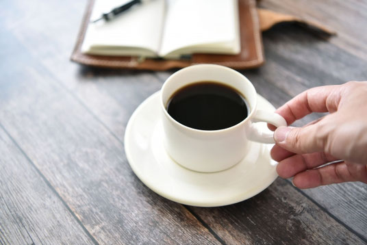 ページがめくられた革のシステム手帳とボールペン。コーヒーの入ったカップ＆ソーサに添えられた手。