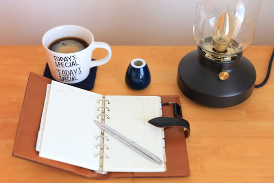 デスクに広げられた茶革のシステム手帳。ボールペン。ペンスタンド。コーヒーの入ったマグカップ。デスクライト。