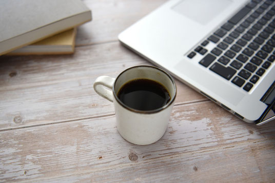 コーヒーの入ったマグカップ。傍らにノートパソコンと積まれた本。