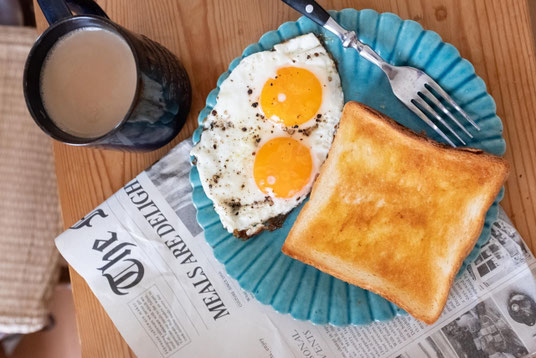 通勤前の食卓。新聞の上に置かれた朝食プレート。目玉焼きとトーストパン。コーヒーの入ったマグカップ。