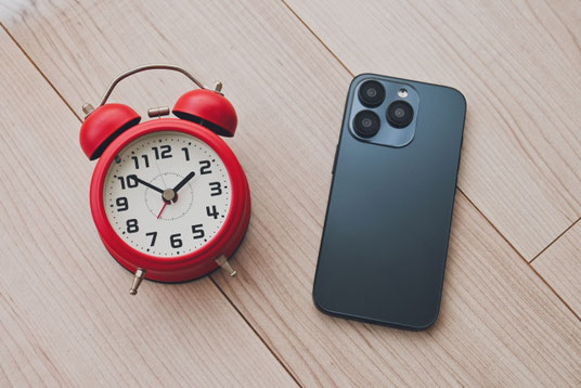木製のテーブルの上に置かれた赤色の目覚まし時計とスマートフォン。