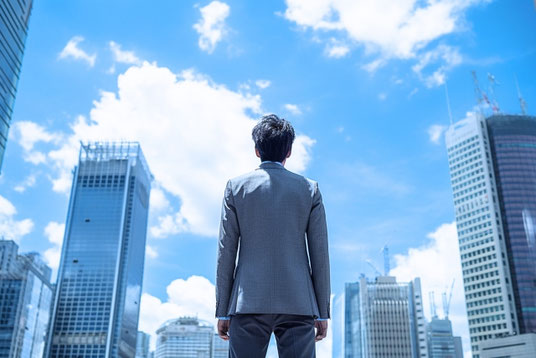 ビルがそびえたつ街並みを眺めるスーツ姿の男性の後ろ姿。