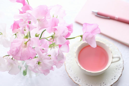スイトピーが生けられた花瓶。紅茶の入ったカップ&ソーサ。ノートとボールペン。