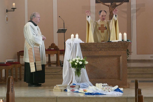 Blick in den Altarraum, in dem vor dem Altar Kerzen unter einem Blumenstrauß niedergelegt sind, die gesegnet werden. Pfarrer Schmitz spricht den Segen, Diakon Thomas Becker assistiert.