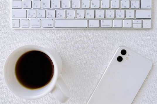 パソコンのキーボードの前に置かれたコーヒーカップとスマートフォン。