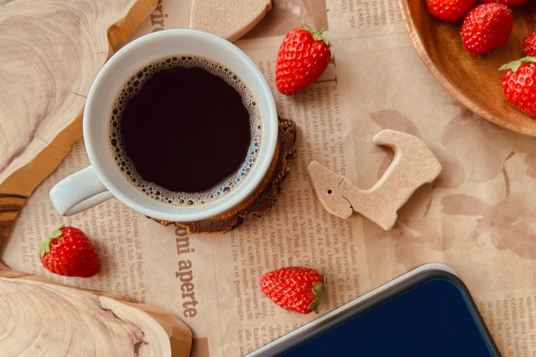 タブロイド紙のうえに置かれたコーヒーの入ったマグカップ、スマートフォン、木製のオーナメント、イチゴが盛り付けられた器。