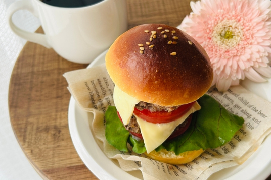 英文資料ペーパーのうえにチーズバーガー。コーヒーの入ったマグカップ。ピンク色のガーベラの花。