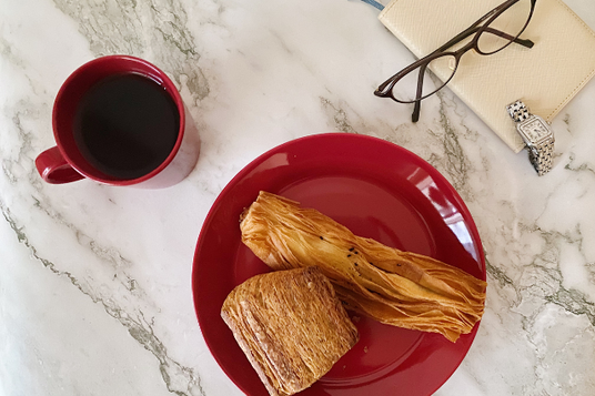 朝食のテーブル。ホットコーヒーのマグカップとデニッシュパンのお皿。手帳の上に置かれた眼鏡と腕時計。通勤。