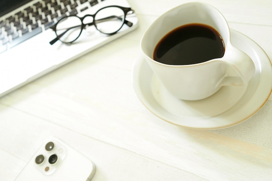 コーヒーの入ったカップ&ソーサ。ノートパソコンのキーボードの上に置かれた黒縁眼鏡。スマートフォン。