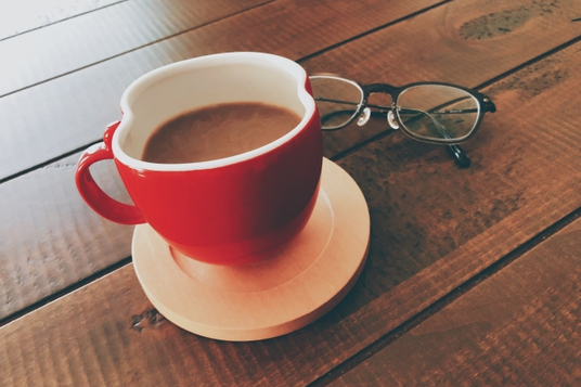 コースターが敷かれたコーヒーのマグカップ。傍らに眼鏡。