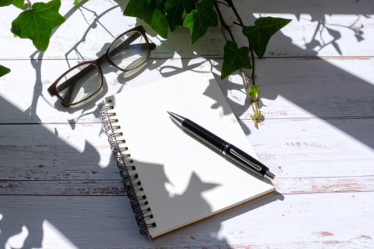 白木のテーブルに置かれたリングノート、ボールペン、眼鏡。傍らにアイビーの葉。