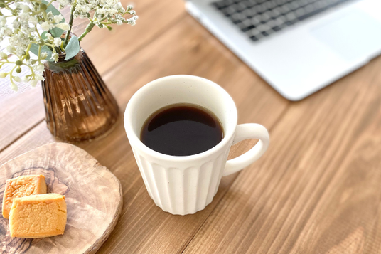 オフィスのデスクに広げられたノートパソコン。傍らにクッキーのお皿、コーヒーのマグカップ、小花が飾られた一輪挿し。