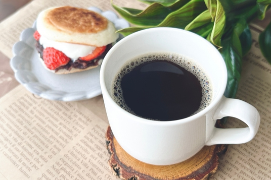 英字新聞のうえにコーヒーの入ったマグカップと生クリームといちごがサンドされた焼き菓子のお皿が置かれている。グリーンの葉。