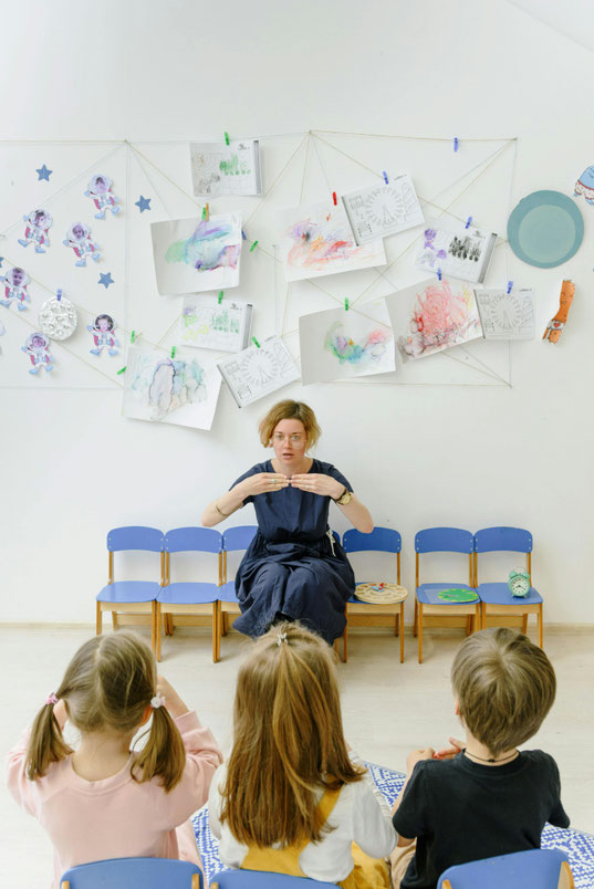 Lehrperson_sitzt_auf_Stuhl_erklaert_drei_Kindern_etwas