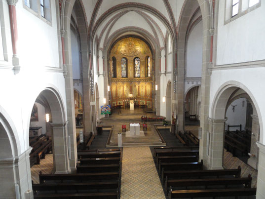 Der Chorraum mit goldenen Gemälden steht im Fokus des Bildes. Vor den braunen Holzkirchenbänken steht der weiße quadratische Altar.