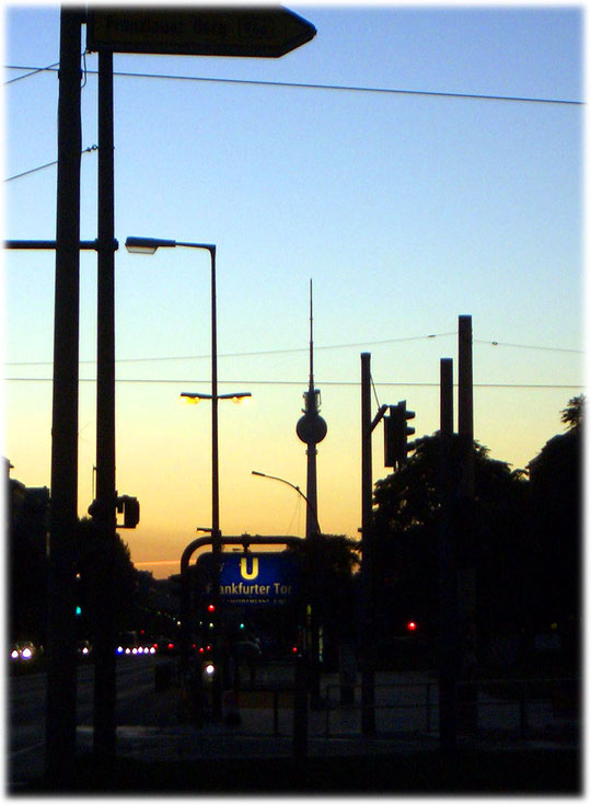 Ein Bild vom Fernsehturm, vom Bezirk Friedrichshain aus fotografiert. Auf dem Foto ist eine Abendstimmung zu sehen, die den Turm in ein schönes warmes Licht taucht. Karl-Marx- und Frankfurter Allee