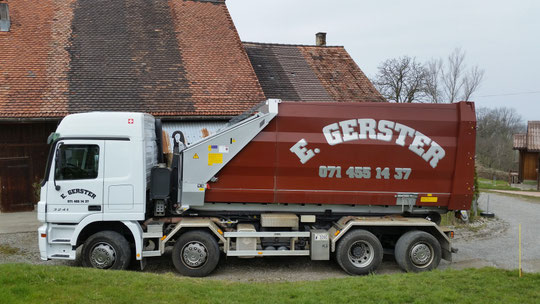 Ein weisser Lastwagen von Gerster Recycling mit Presscontainer und Pressmulde. Im Hintergrund sind mehrere grosse rote Abfallcontainer mit dem Logo von Gerster Recycling zu sehen. Das Bild zeigt einen Recycling- und Abfallentsorgungsbetrieb.