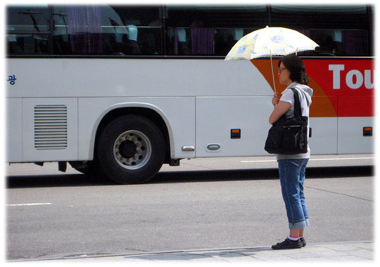 This image shows a young woman standing at a bus station and protecting her body with a sunshade. She doesn't wants to get tanned! Fotos von koreanischen Frauen die in unter einem Sonnenschirm steht