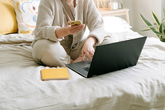 自宅でのプライベートタイムに電話対応。ベッドでスマートフォンを操作しながらノートパソコンと手帳を広げる女性。
