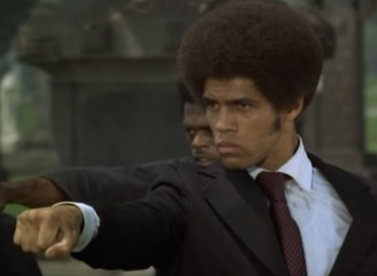 Auf dem Bild ist Jim Kelly in einem schwarzen Herrenanzug zu sehen. Man sieht nur den Oberkörper. Er führt einen Karateschlag aus.