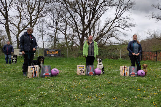 Drei Frauen mit ihren Hunden stehen auf einer Wiese vor Bäumen und halten Preise mit nummerierten Podesttafeln.