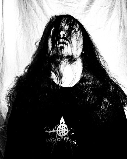 Black and white photo of a young man with a short beard and long hair falling into his face, looking up. He is wearing a black sweatshirt with a white print from the streetwear brand DAYS OF CHAOS™. The design shows a burning globe, a sword and lightning.
