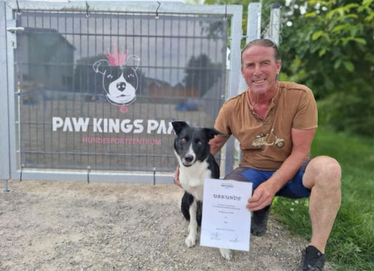 Wolfgang kniet neben schwarz-weißem Hund Boh und hält eine Urkunde vor einem Zaun mit Logo und Schriftzug 'PAW KINGS PARK HUNDESPORTZENTRUM'.