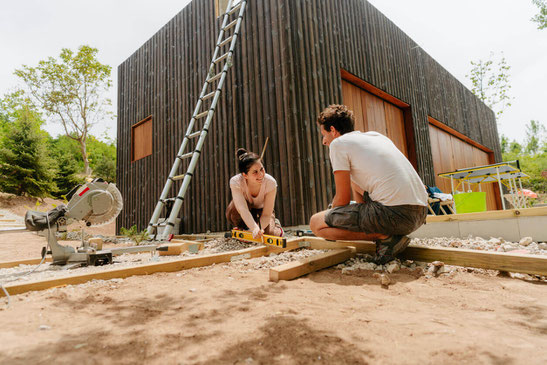 Ein Paar beim Verlegen der Unterkonstruktion für eine Bambusterrasse vor einem modernen Haus mit dunkler Holzfassade – handwerkliche Arbeiten im Garten bei sonnigem Wetter