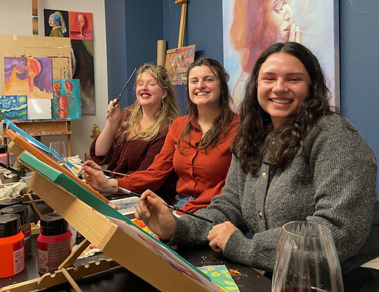 Lachende deelnemers tijdens een gezellige Sip & Paint-workshop bij Daphne Schrijver in Breda, omringd door verf, doeken en kleurrijke schilderijen.