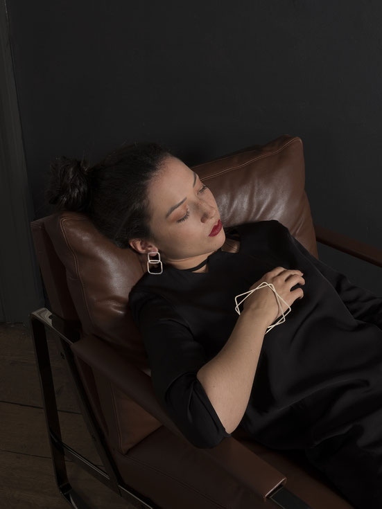 jeune femme allongée dans un fauteuil en cuir portant une grande boucle d'oreilles composée de deux carrés en argent et un bracelet en argent