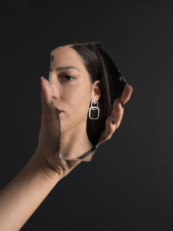 main de femme qui tient un miroir dans lequel on voit un visage portant une bocule d'oreilles composée de deux carrés en argent