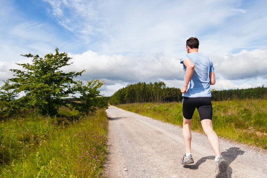 Ein Mann in Sportkleidung joggt im Freien auf einem Gehweg zwischen grüner Wiese und mit einem Wald im Hintergrund.