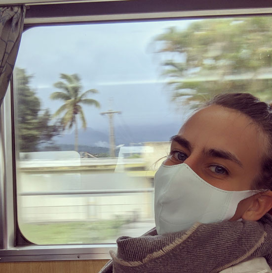 Portrait of Miss Vizzz with a Facemask in Taiwanese Train, passing through the landscape. Through the window of the train behind the artist you can se palms, houses and mountains.