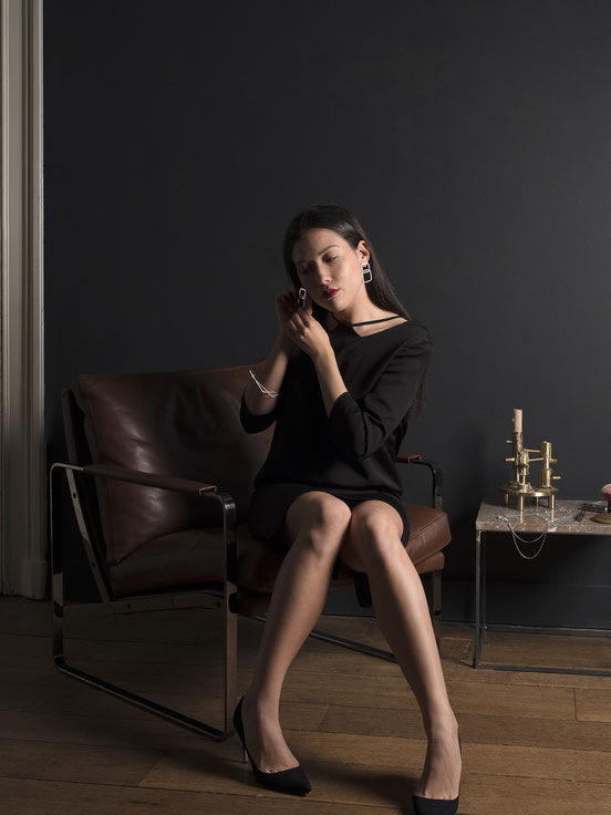 jeune femme qui s'apprête assise dans un fauteuil en cuir et se met des boucles d'oreilles composées de deux carrés en argent