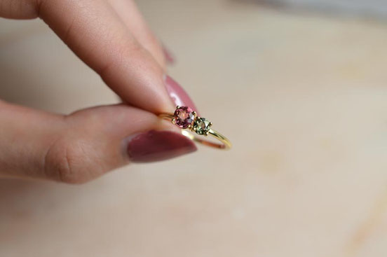 Main de femme qui tient une bague de fiançailles en or jaune recyclé sertie de deux tourmalines verte et rose