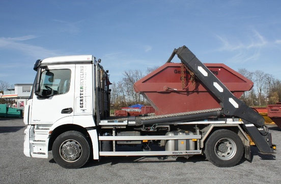 Ein weisser Lastwagen von Gerster Recycling mit Mulde, Container, Box. Im Hintergrund sind mehrere grosse rote Abfallcontainer mit dem Logo von Gerster Recycling zu sehen. Das Bild zeigt einen Recycling- und Abfallentsorgungsbetrieb.