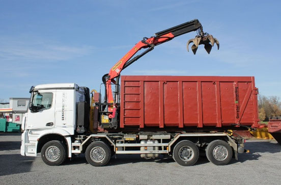Ein weisser Lastwagen von Gerster Recycling mit Kran und Greifer. Im Hintergrund sind mehrere grosse rote Abfallcontainer mit dem Logo von Gerster Recycling zu sehen. Das Bild zeigt einen Recycling- und Abfallentsorgungsbetrieb.